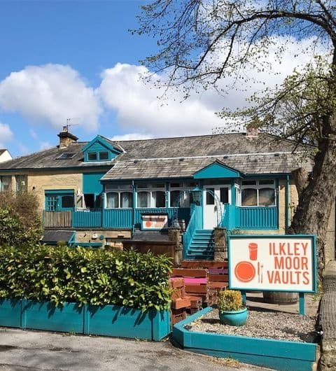 Exterior view of Ilkley Moor Vaults with blue-trimmed windows, a signboard, and surrounding greenery on a sunny day.