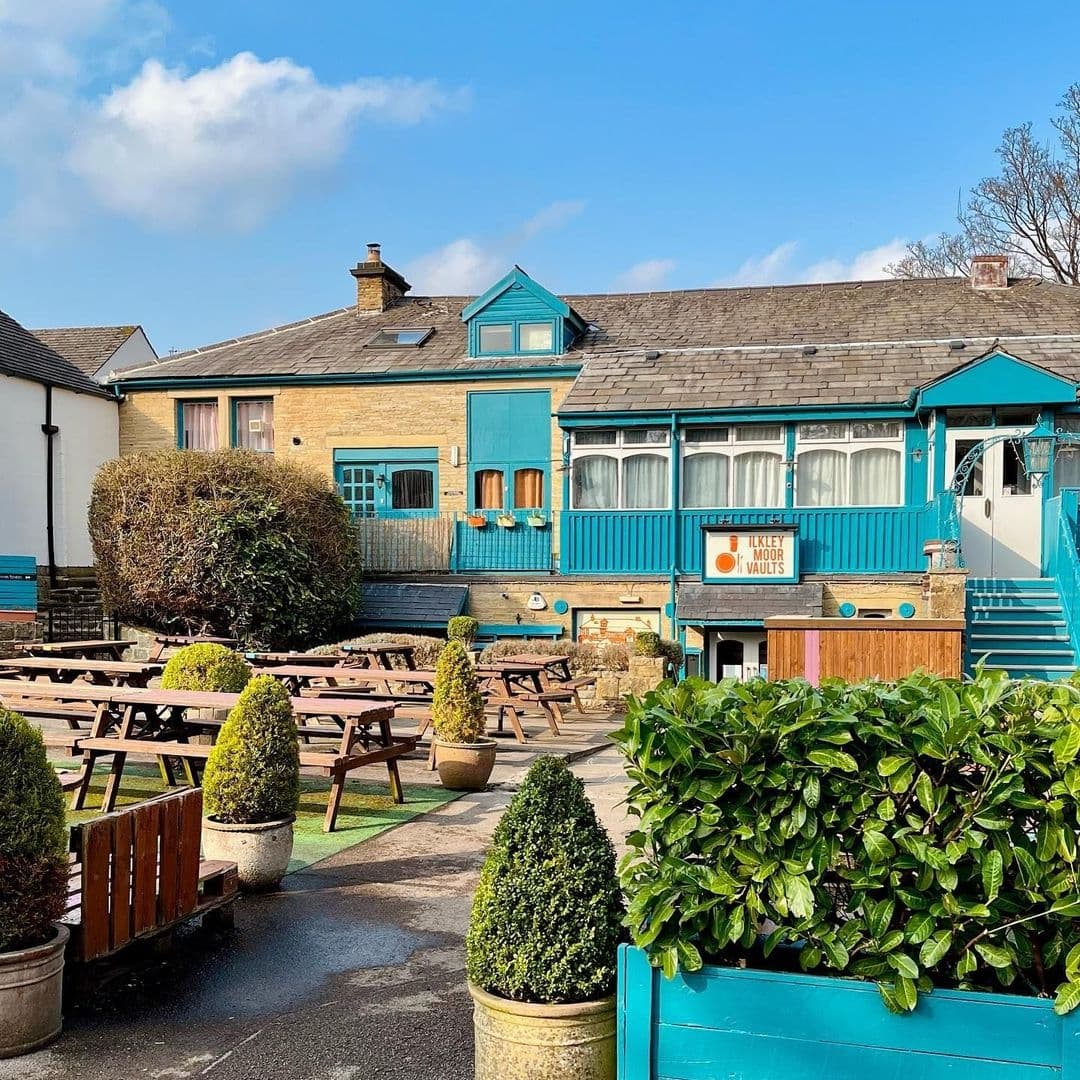 Exterior view of Ilkley Moor Vaults with outdoor wooden picnic tables, topiary in planters, and a blue-trimmed building facade under a clear sky.