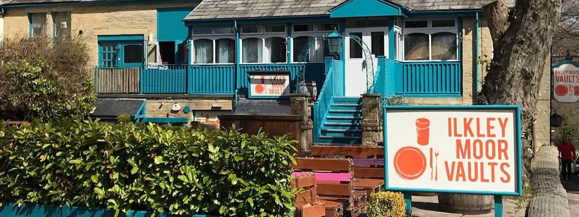 Exterior view of Ilkley Moor Vaults featuring a stone building with bright blue trim, a front patio with wooden planters, and a sign displaying the establishment's name.