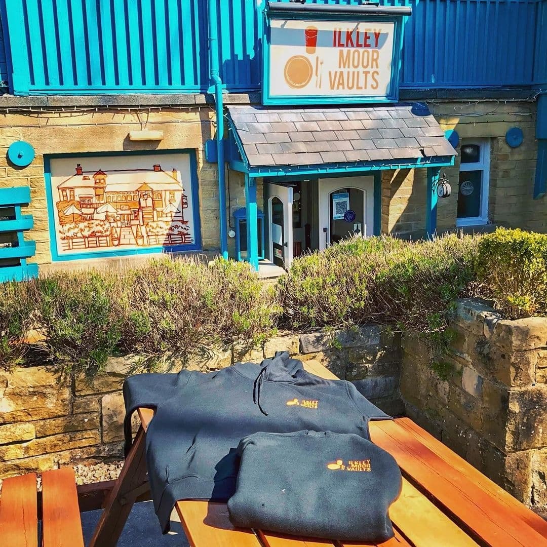 Exterior view of Ilkley Moor Vaults with a blue sign above the entrance, a stone building with blue trim, and a mural depicting a historic scene. A wooden table in the foreground displays black hoodies with the establishment's logo.
