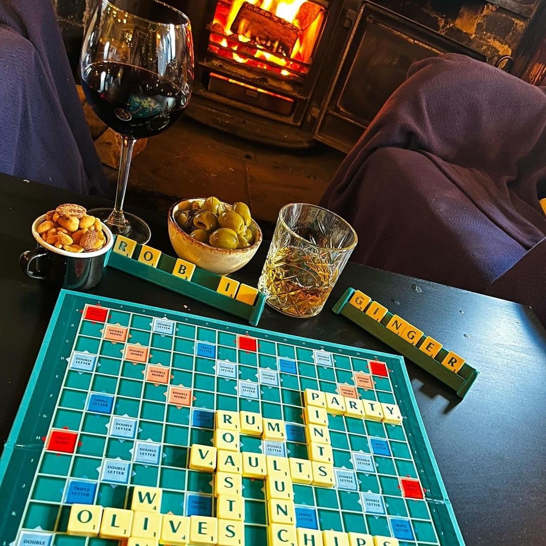 Cozy atmosphere at Ilkley Moor Vaults with a Scrabble board game on a table, a glass of red wine, a tumbler of whiskey, bowls of nuts and olives, and a roaring fireplace in the background.