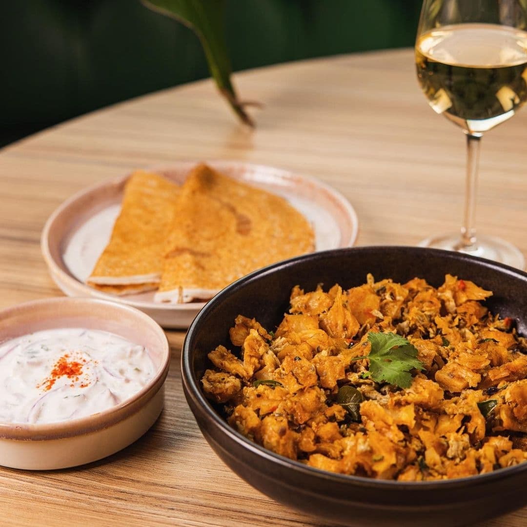 Traditional Indian cuisine at Ma-Hé Coastal Indian featuring a bowl of spicy chicken curry garnished with coriander, a side of flatbread, a small bowl of raita, and a glass of white wine on a wooden table.