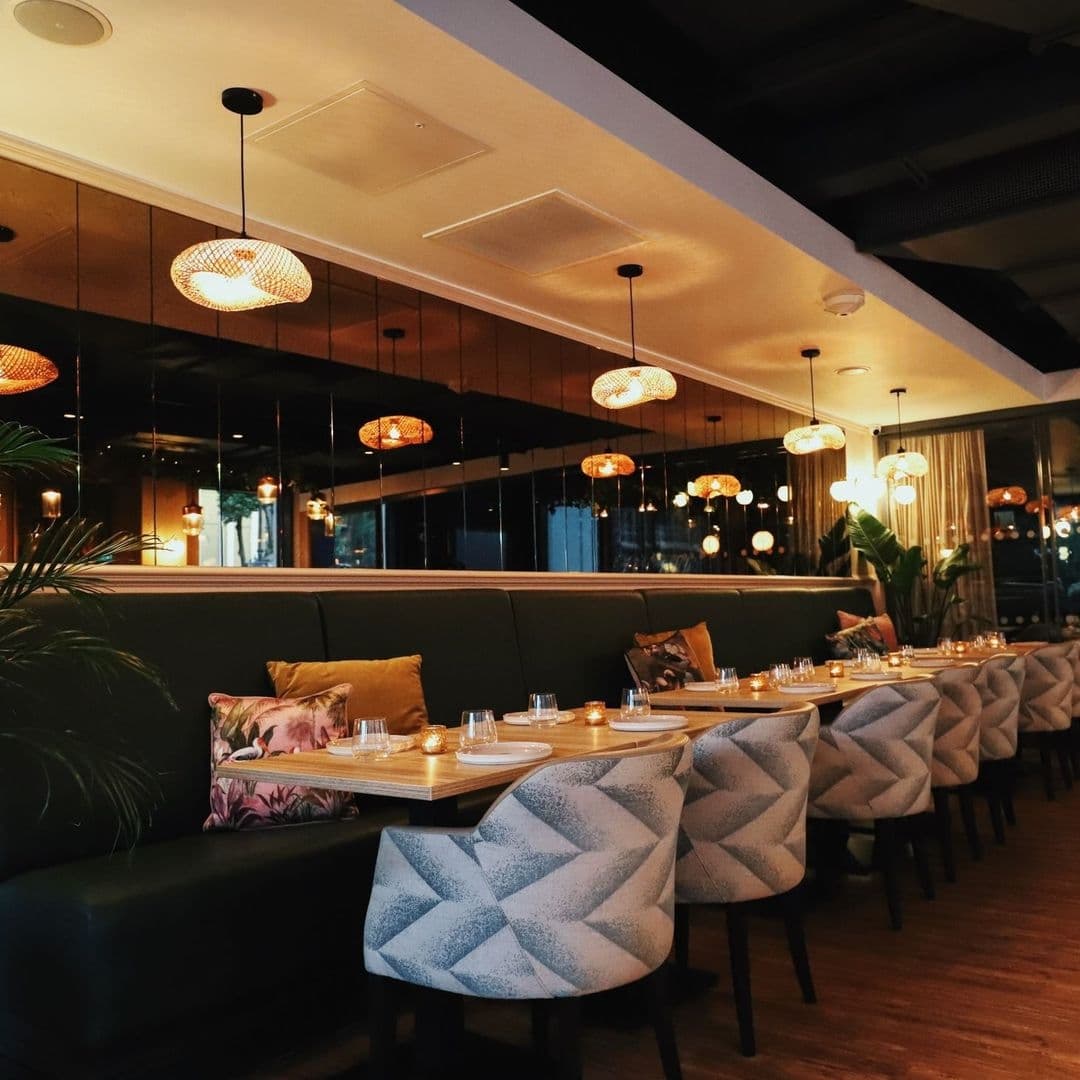 Elegant dining area at Ma-Hé Coastal Indian with patterned chairs, green booth seating, decorative pendant lights, and tables set with glassware.