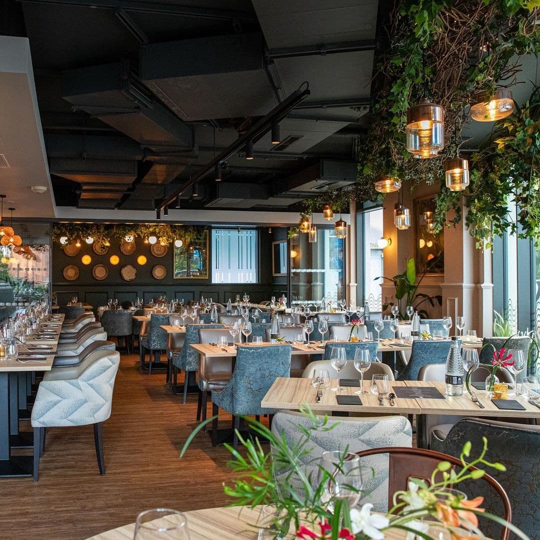 Elegant dining area at Ma-Hé Coastal Indian, featuring neatly arranged tables with wine glasses, hanging greenery, and warm lighting.