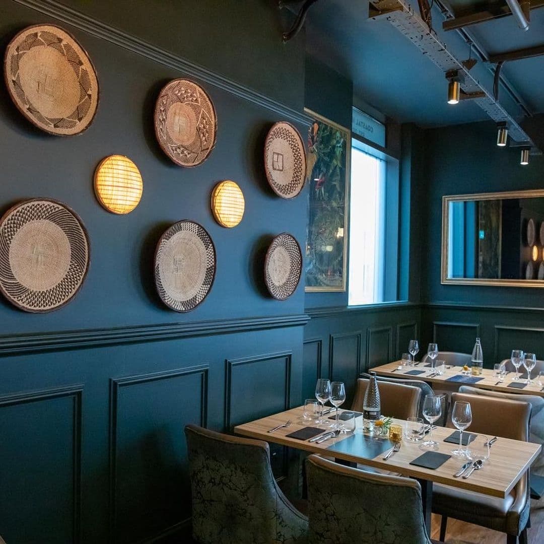 Elegant dining room at Ma-Hé Coastal Indian featuring teal walls adorned with decorative woven plates, warm lighting, and sophisticated table settings.