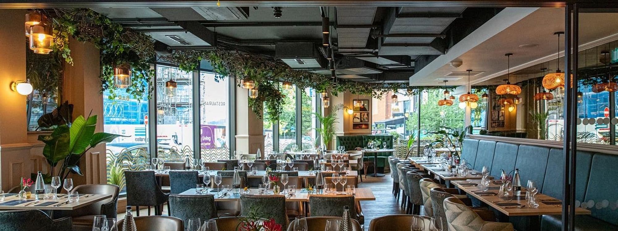 Interior of Ma-Hé Coastal Indian restaurant showing elegantly set tables with wine glasses, pendant lighting, greenery, and a view of the street outside.