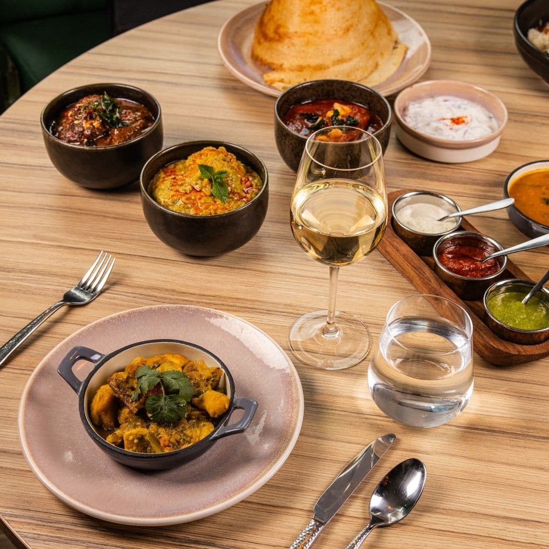 Assorted traditional Indian dishes and condiments served at Ma-Hé Coastal Indian, featuring curries, chutneys, a fluffy bread, with a glass of white wine and water on a wooden table.