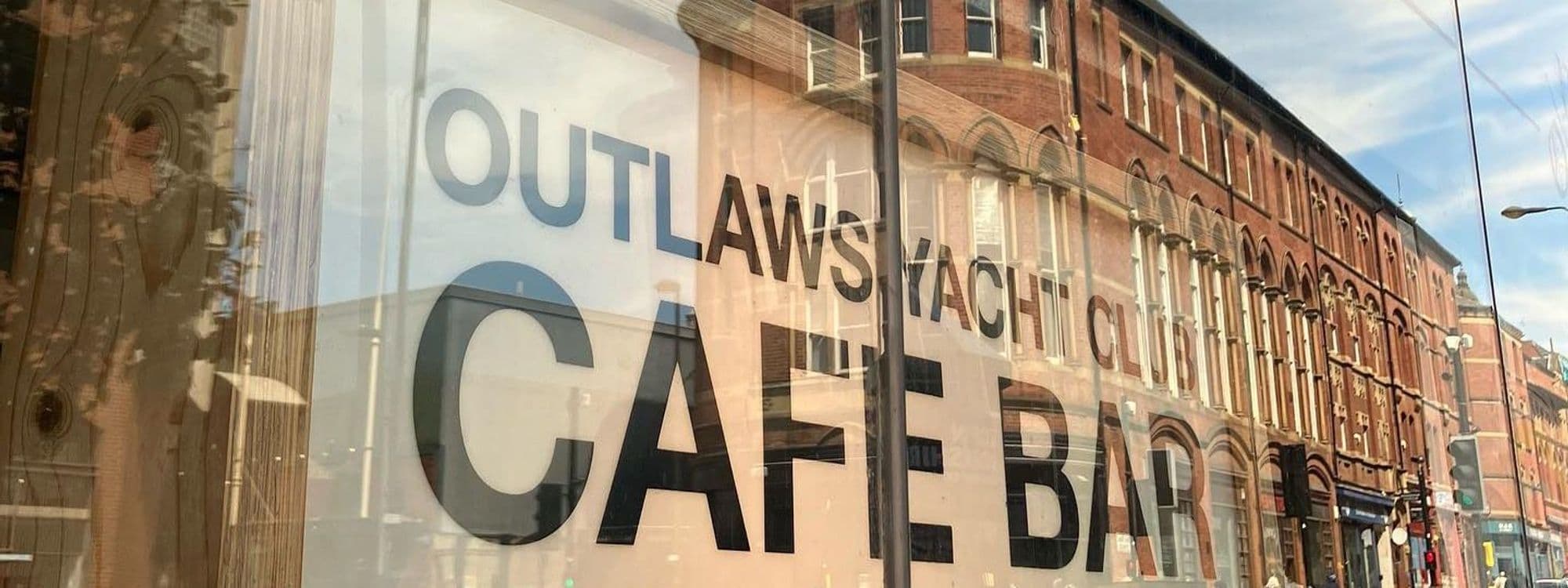 Exterior view of Outlaws Yacht Club with reflective window showcasing the cafe bar's signage and a brick building facade in the background.