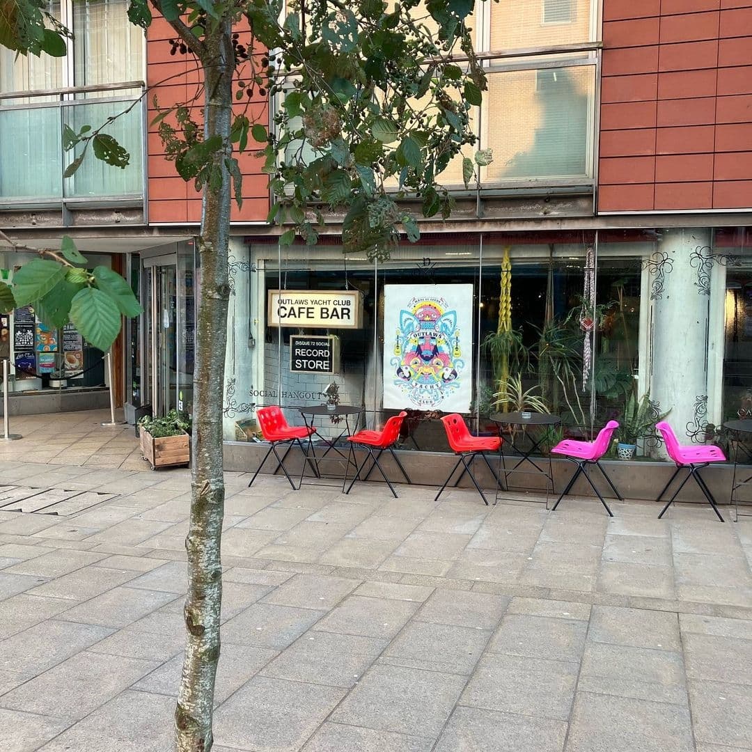 Exterior view of Outlaws Yacht Club with vibrant red and pink chairs lined up outside, a tree in the foreground, and large windows displaying posters and greenery.