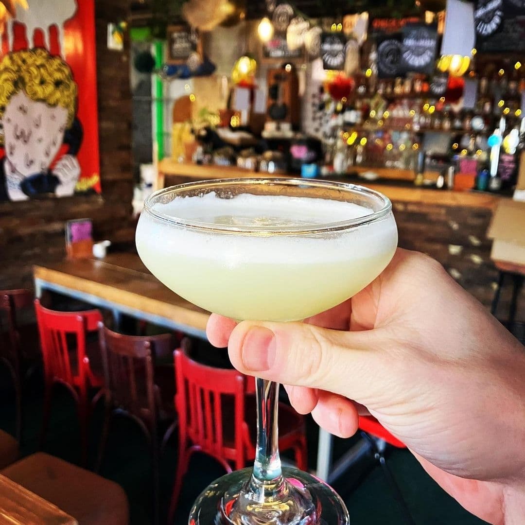 Hand holding a cocktail at Outlaws Yacht Club with the bar's cozy interior and eclectic decor in the background.