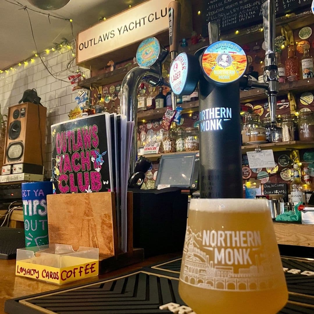 Inside Outlaws Yacht Club, with a close-up of a beer tap labeled 'Northern Monk' and a glass of beer in the foreground, menus and loyalty cards on the counter, and a variety of spirits on shelves in the background.