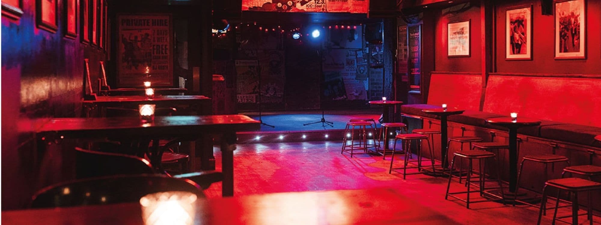 Interior of Smokestack featuring dim lighting with red and blue hues, wooden tables, stools, and a stage set for live performances, surrounded by walls adorned with posters and artwork.