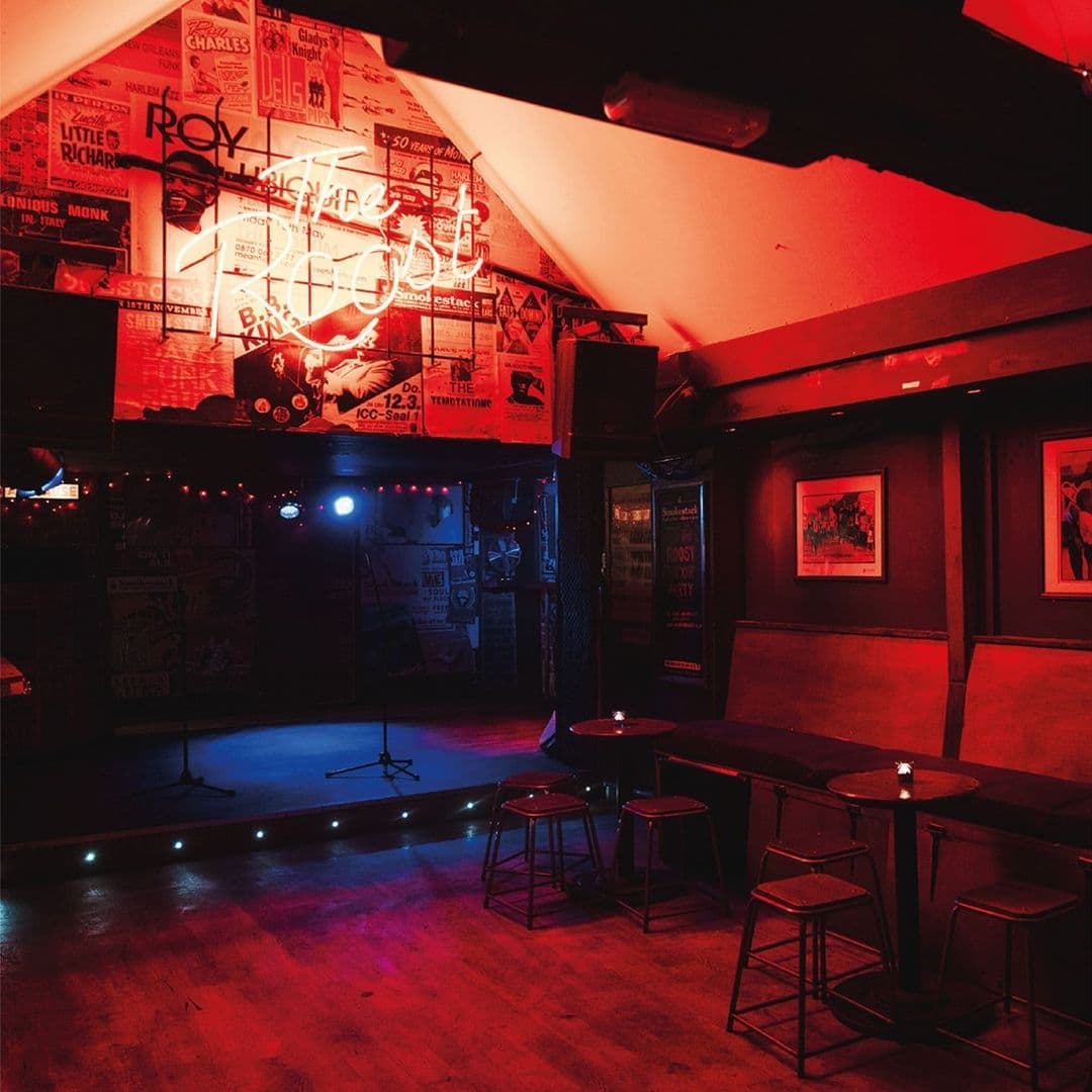 Interior of Smokestack featuring dim red lighting, a stage with a microphone stand, bar stools, and walls adorned with music posters and neon signs.