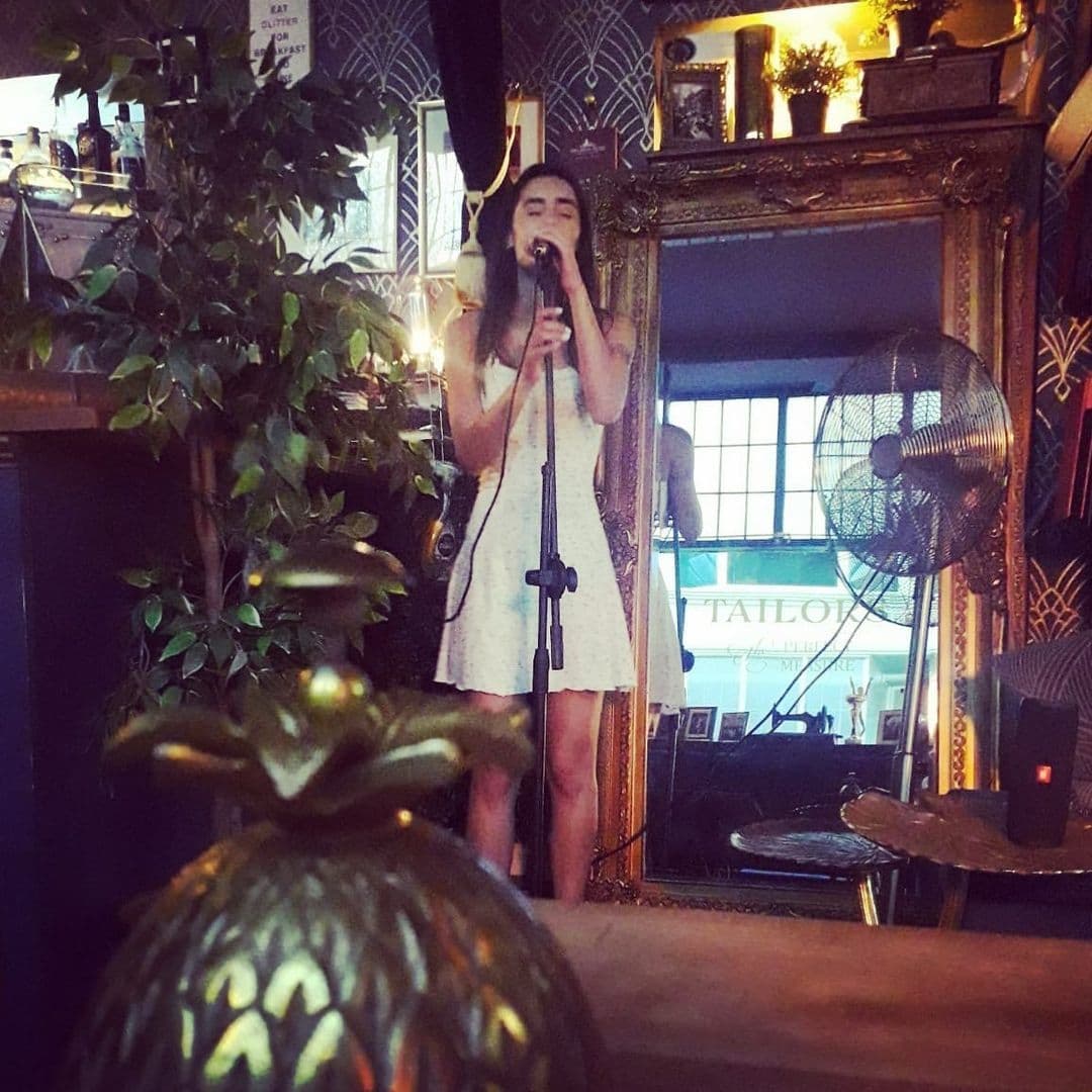 Woman singing into a microphone at Tailors Gin, reflected in a large framed mirror surrounded by ornate decor and green plants.