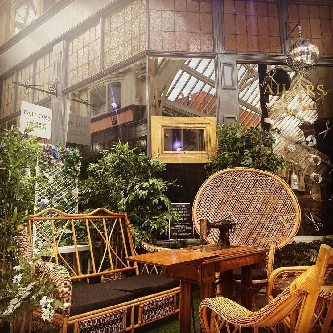 Cozy outdoor seating area at Tailors Gin with rattan chairs, lush greenery, and vintage decor under a glass roof.