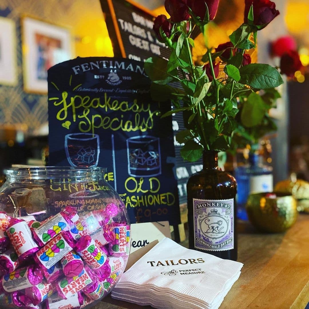 A cozy bar scene at Tailors Gin featuring a vase with red roses, a jar of pink wrapped candies, a bottle of Monkey 47 Schwarzwald Dry Gin, and a napkin with the Tailors logo. In the background, a chalkboard displays 'Speakeasy Specials' with cocktail offers.