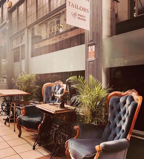 Outdoor seating at Tailors Gin with elegant vintage-style blue tufted chairs and wooden tables, flanked by green potted plants, in front of the establishment's window displaying the Tailors sign.