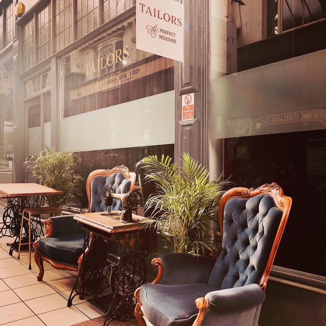 Outdoor seating area at Tailors Gin with vintage-style upholstered chairs and sewing machine tables, surrounded by potted plants, under the warm glow of sunlight.