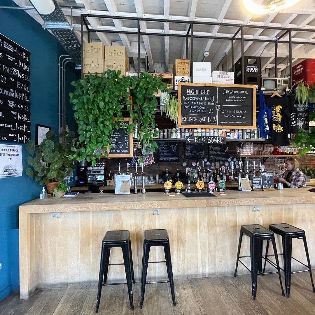 Interior view of The Brunswick featuring a well-stocked bar with beer taps, blackboard menus, wooden bar stools, and green potted plants.