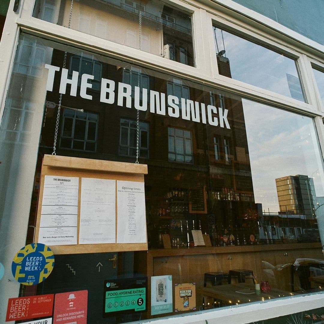 Exterior view of The Brunswick showing a large window with the name in bold letters, menus on display, and various event stickers, reflecting the vibrant city life outside.