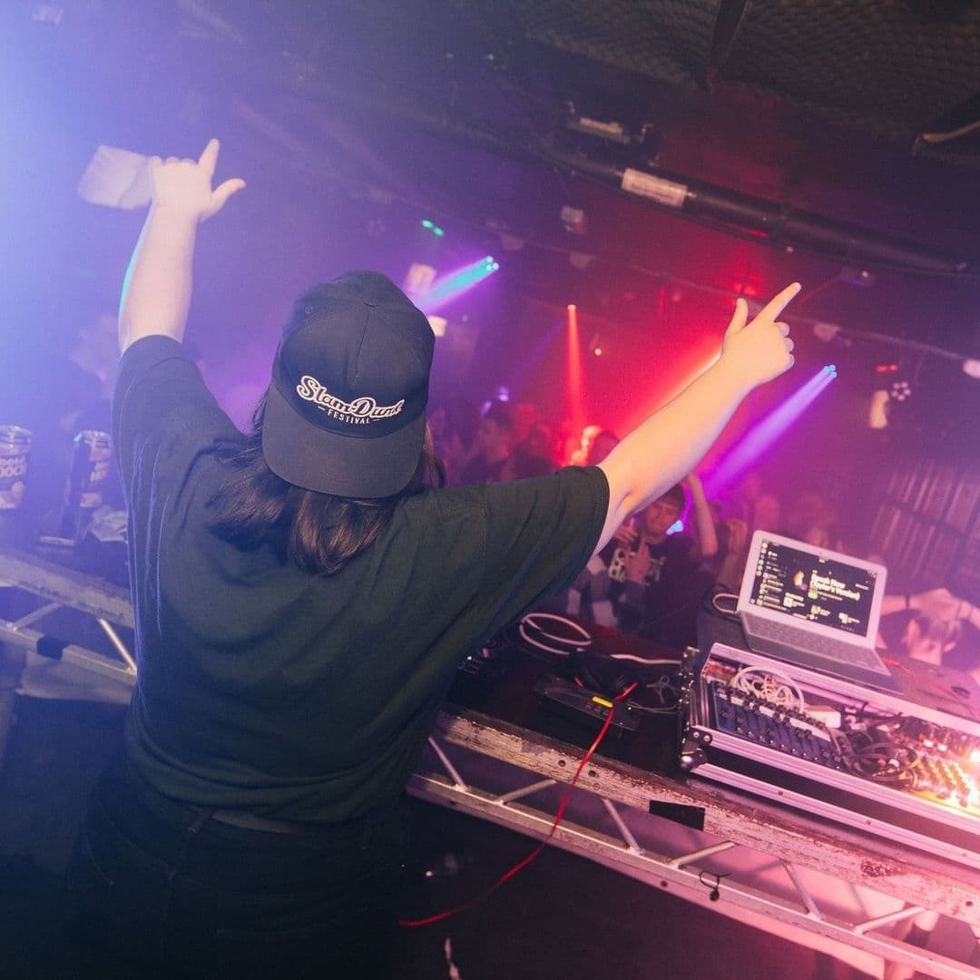 DJ performing at The Key Club with hands up and crowd in the background under colorful lights.