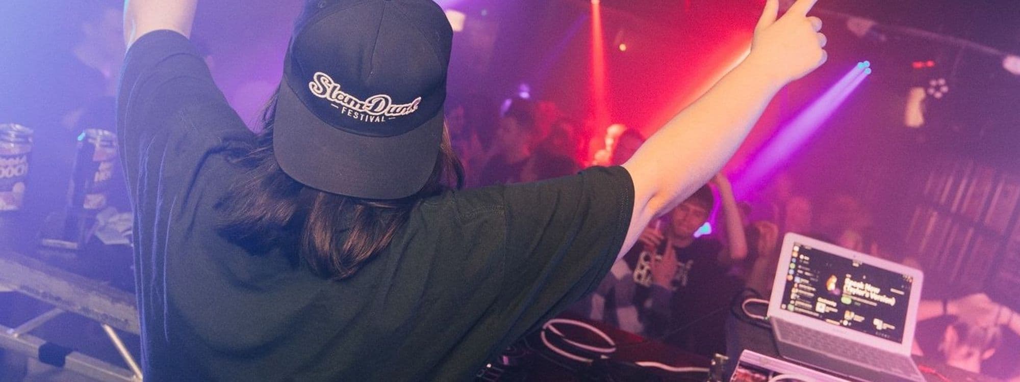 DJ performing at The Key Club with vibrant purple lighting, raising hand to the crowd, with a laptop and sound equipment on the table.