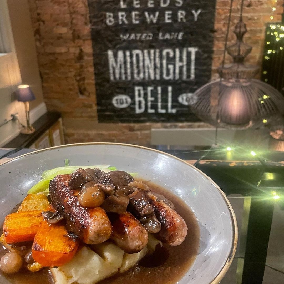A hearty meal of sausages with mashed potatoes and gravy served on a plate, with a backdrop featuring The Midnight Bell sign from Leeds Brewery.