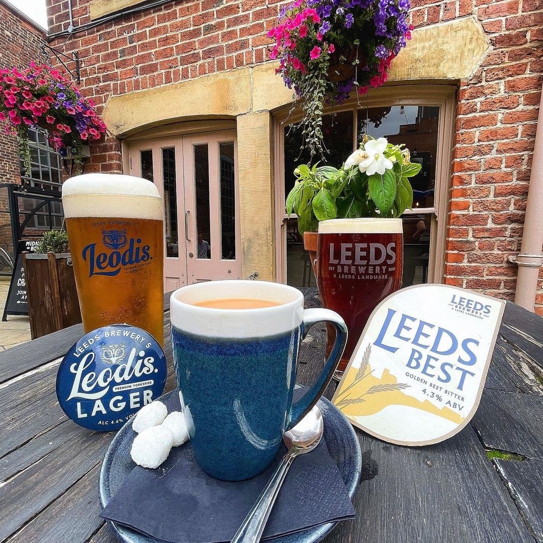 Outdoor table at The Midnight Bell with a pint of Leodis Lager, a cup of tea, and a Leeds Best beer mat, with a backdrop of a brick building and hanging flower baskets.