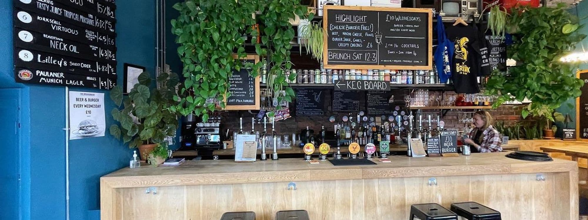 Interior of The Brunswick featuring a bar with a variety of taps and a chalkboard menu, surrounded by green potted plants and a patron sitting at the end of the bar.