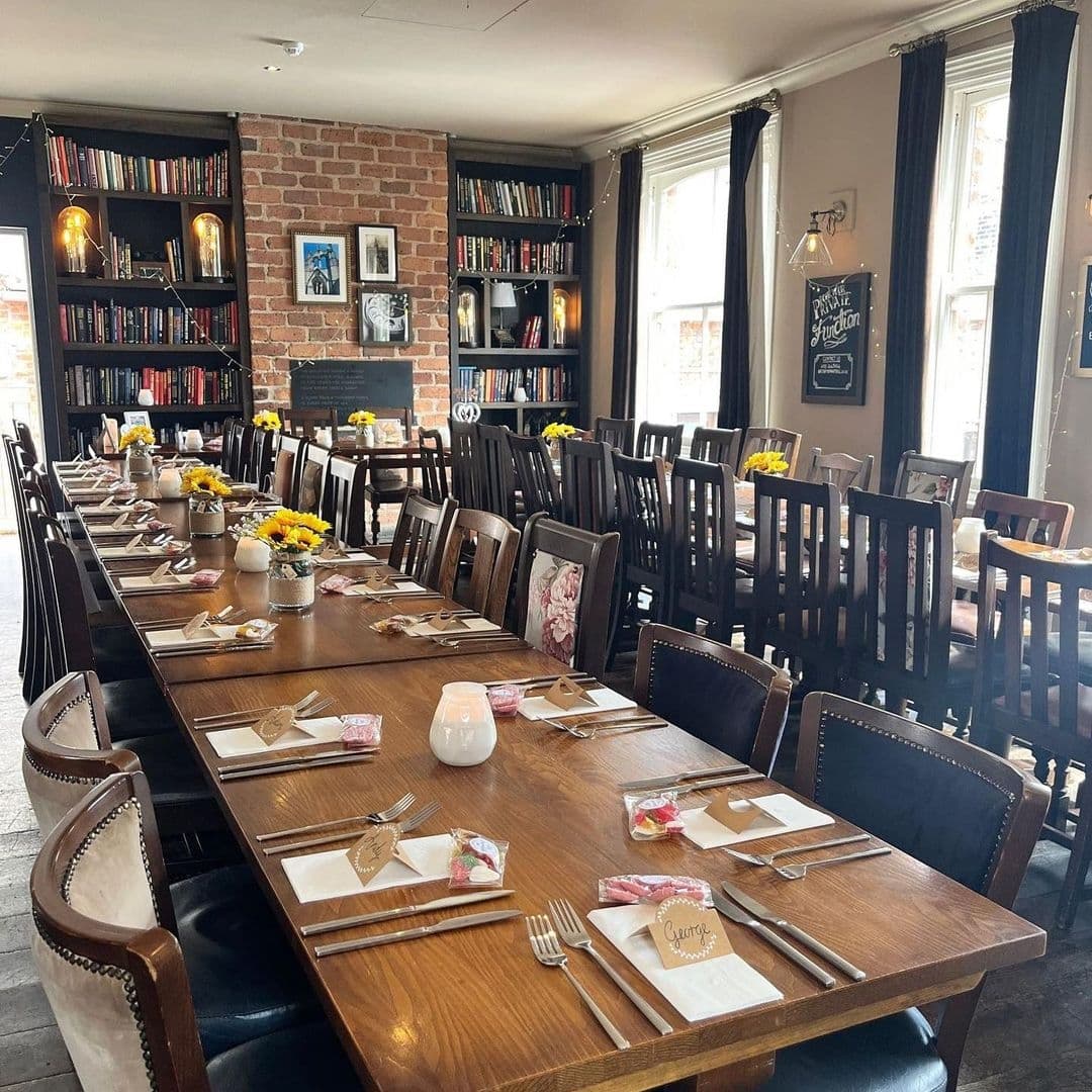 Cozy dining area at The Midnight Bell with wooden tables set for a meal, surrounded by bookshelves, exposed brick walls, and dark curtains.