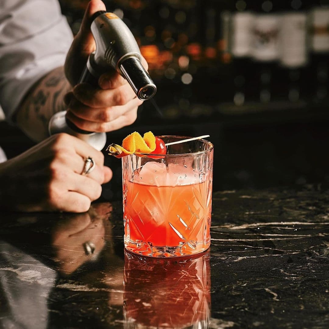 Bartender preparing a cocktail at Blue Pavilion, featuring a glass with a pink beverage garnished with an orange peel on a dark bar top.