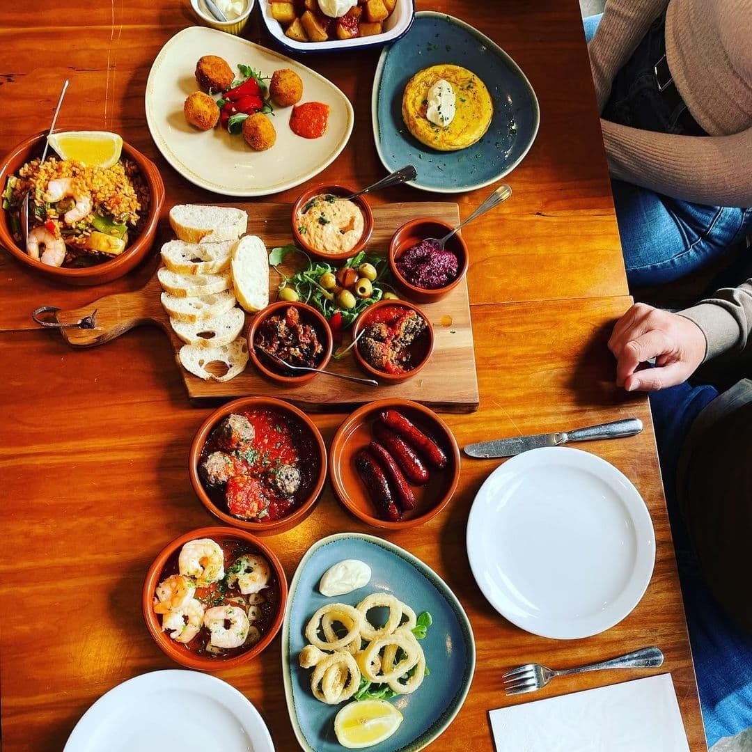 Assorted traditional Spanish tapas and paella at Bomba Paella, featuring dishes like shrimp, chorizo, calamari, meatballs, and croquettes on a wooden table with empty plates and cutlery, ready for dining.