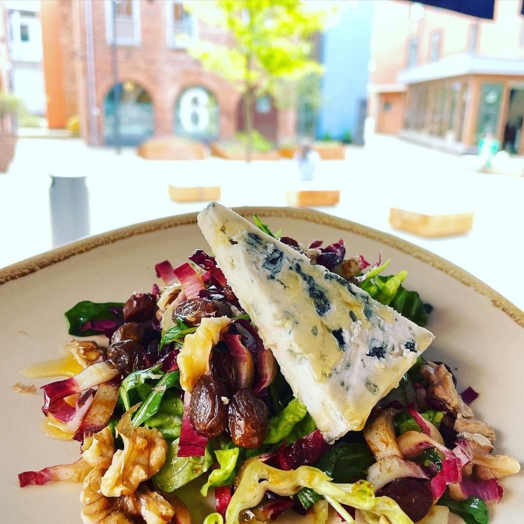 Alt text: "Fresh gourmet salad with blue cheese, walnuts, and raisins served on a plate at Bomba Paella, with a blurred background featuring outdoor seating and a brick building."