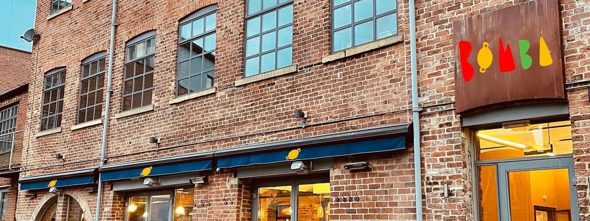 Exterior view of Bomba Paella restaurant with a colorful logo above the entrance, featuring a brick facade and large windows under a blue awning.