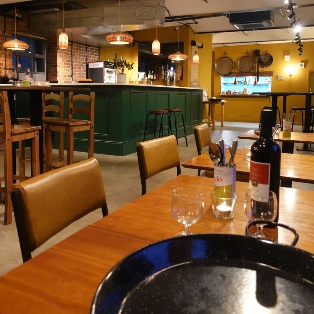 Interior of Bomba Paella restaurant with wooden tables and chairs, a bottle of red wine, and a cozy bar area with pendant lighting.