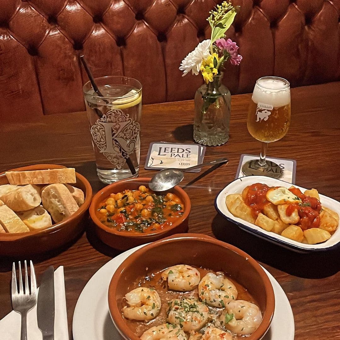 A variety of Spanish dishes and drinks on a wooden table at Bomba Paella, featuring bowls of shrimp, stew, and patatas bravas, with a glass of water and a pint of Leeds Pale beer, complemented by fresh bread and a small vase with flowers.