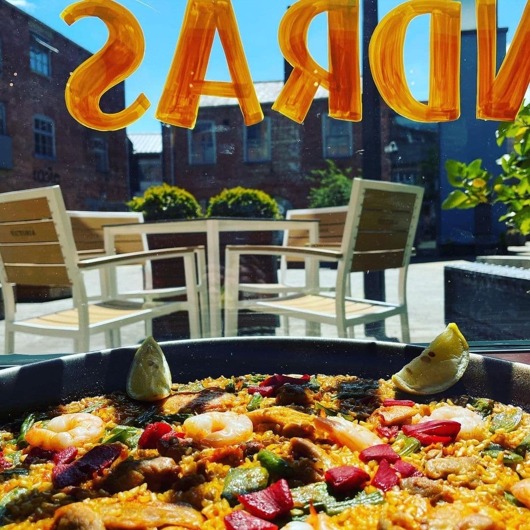 Alt text: "Sunny outdoor dining at Bomba Paella with a close-up of a delicious paella dish, featuring shrimp, vegetables, and slices of lemon, set against a backdrop of outdoor tables and chairs."