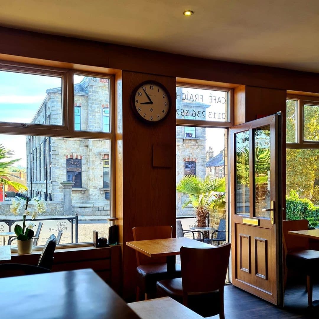 Interior view of Cafe Fraiche with wooden tables and chairs, a wall clock, and large windows offering a sunny street view.