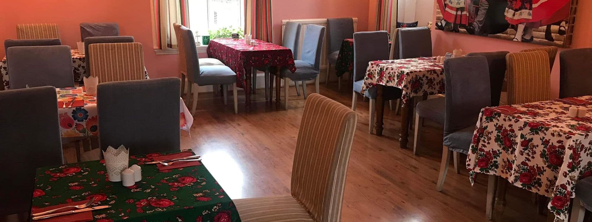 Interior of Cracovia Taste of Poland restaurant showing a cozy dining area with wooden floors, multiple tables set with colorful traditional Polish floral tablecloths, and a variety of upholstered chairs. Bright natural light filters through the windows.