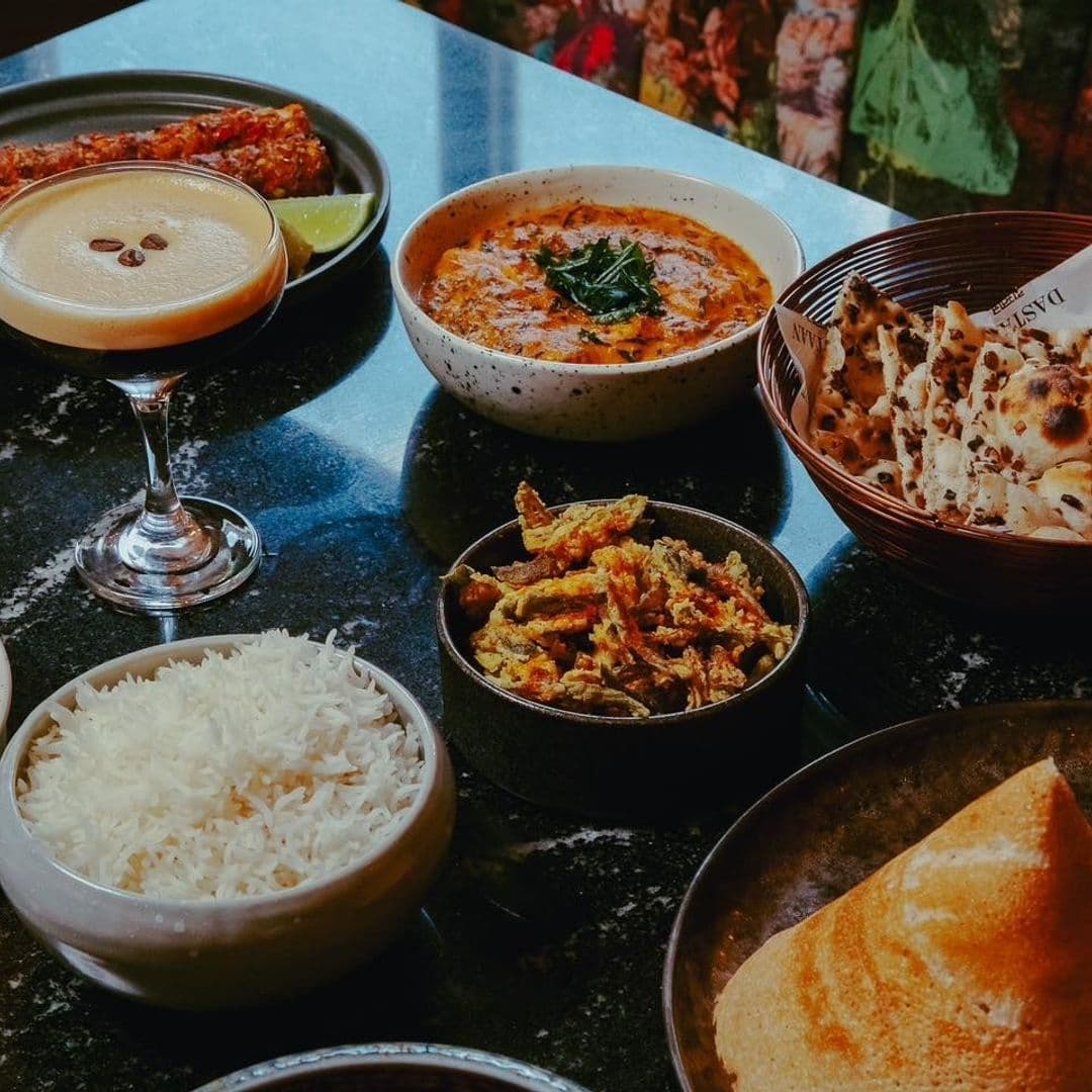 Assorted traditional dishes at Dastaan featuring a creamy cocktail, a bowl of curry with a leaf garnish, basmati rice, crispy flatbread, and a dish of seasoned vegetables.