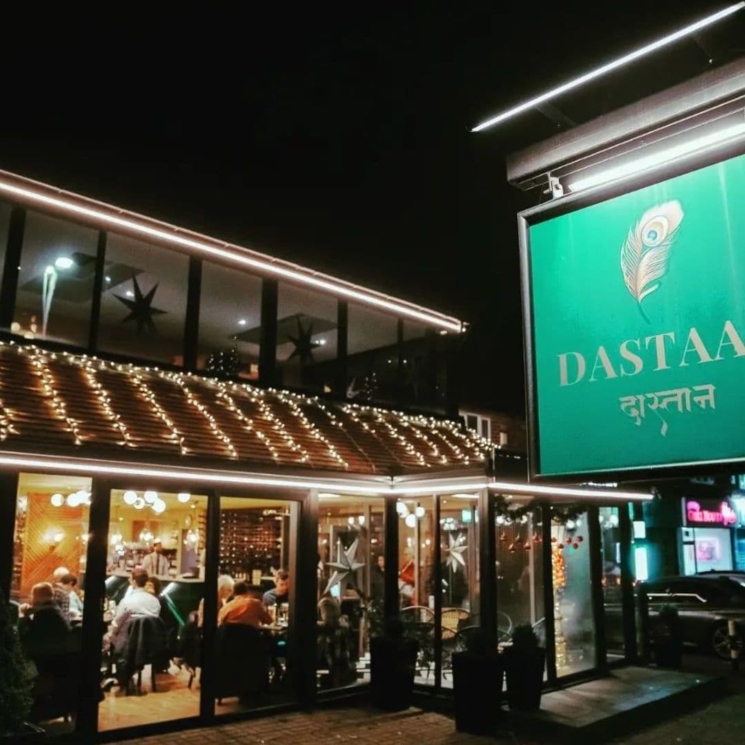 Exterior view of Dastaan restaurant at night with illuminated signboard, string lights on the roof, and visible diners inside through the glass windows.