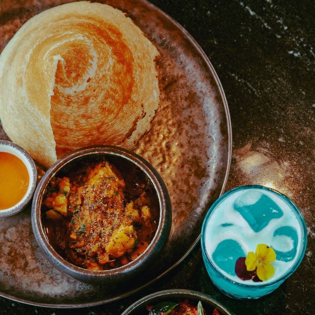 Traditional South Asian meal at Dastaan featuring a crispy dosa, a bowl of spiced potatoes, a side of orange sauce, and a blue-colored drink garnished with a yellow flower.
