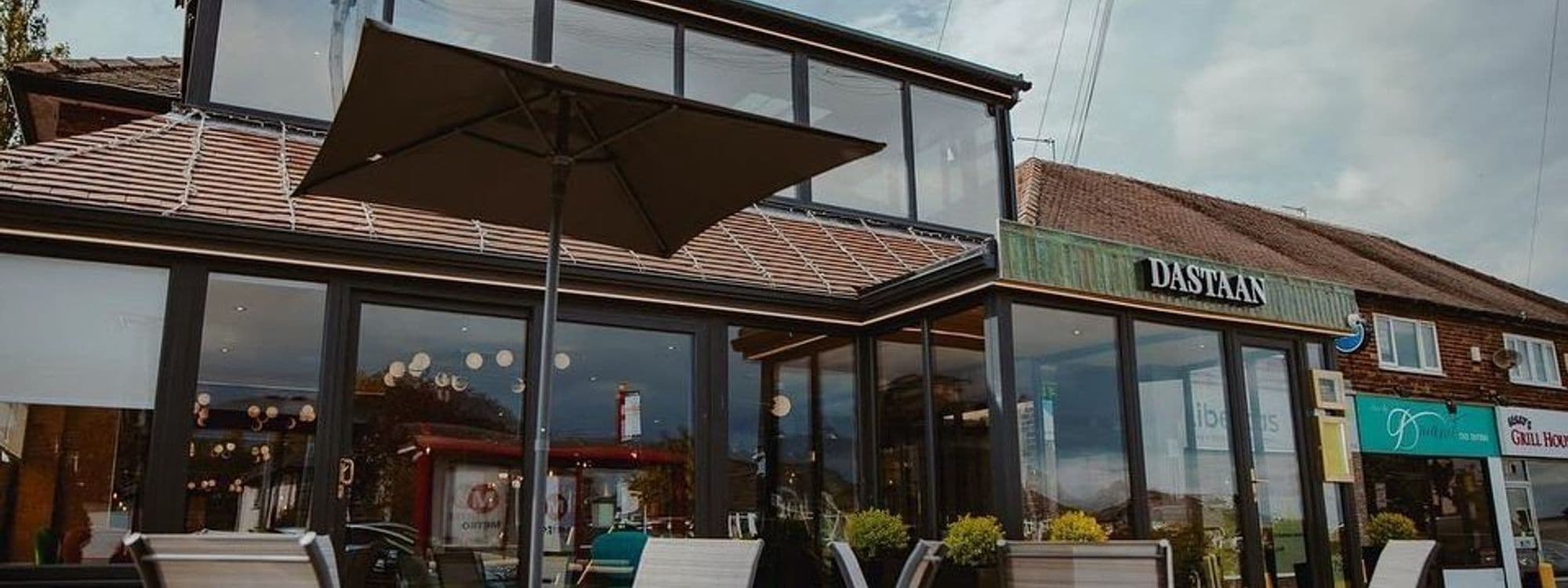 Exterior view of Dastaan with clear glass windows, a tiled roof, and an outdoor seating area shaded by a large umbrella.