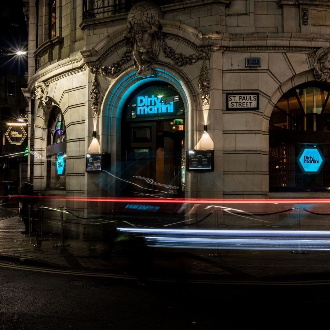 Nighttime view of the Dirty Martini bar entrance with illuminated signage, set in an ornate building on St. Paul's Street, featuring light trails from passing traffic.
