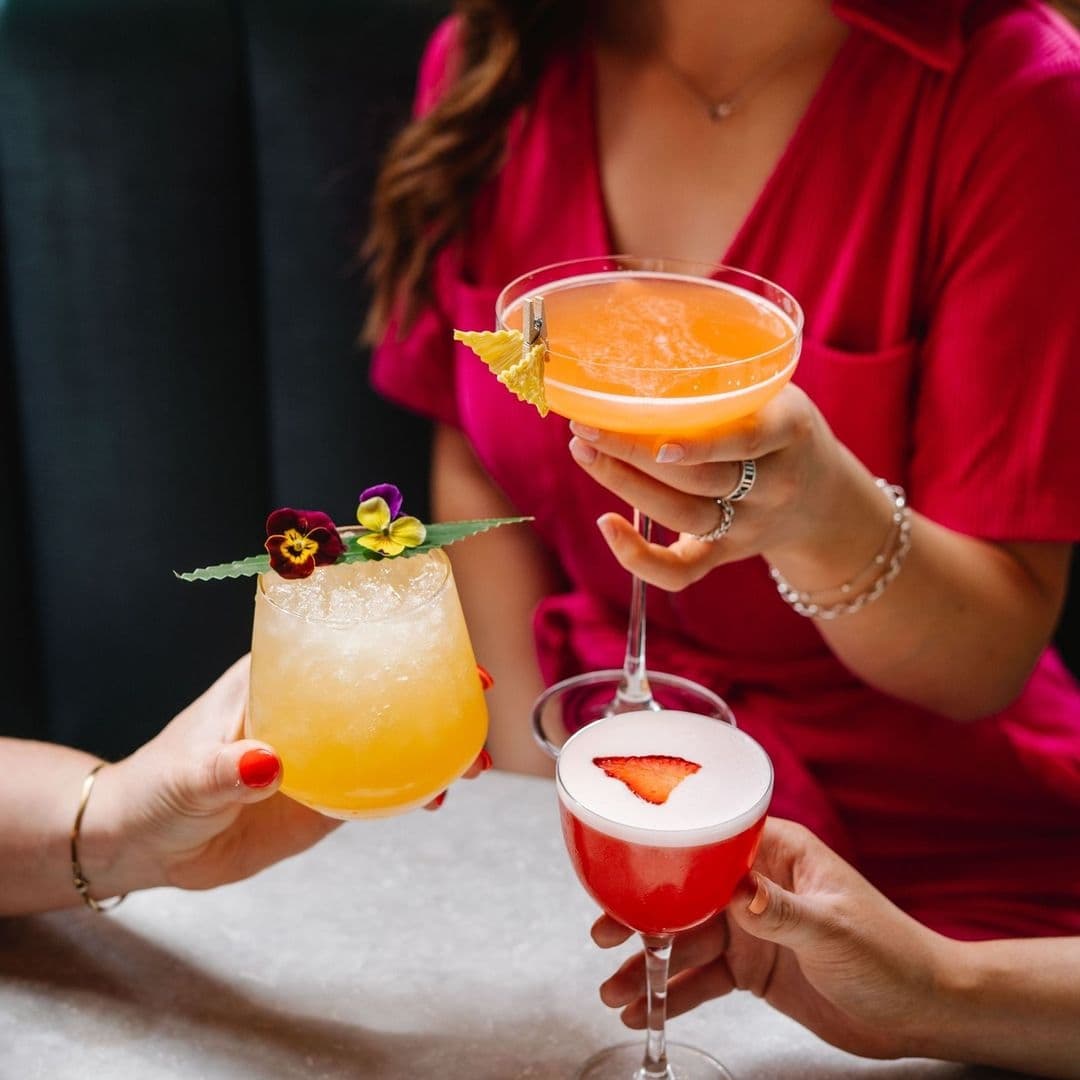 Three colorful cocktails being held by people at Dirty Martini, with a focus on the drinks and a glimpse of a person in a red outfit in the background.