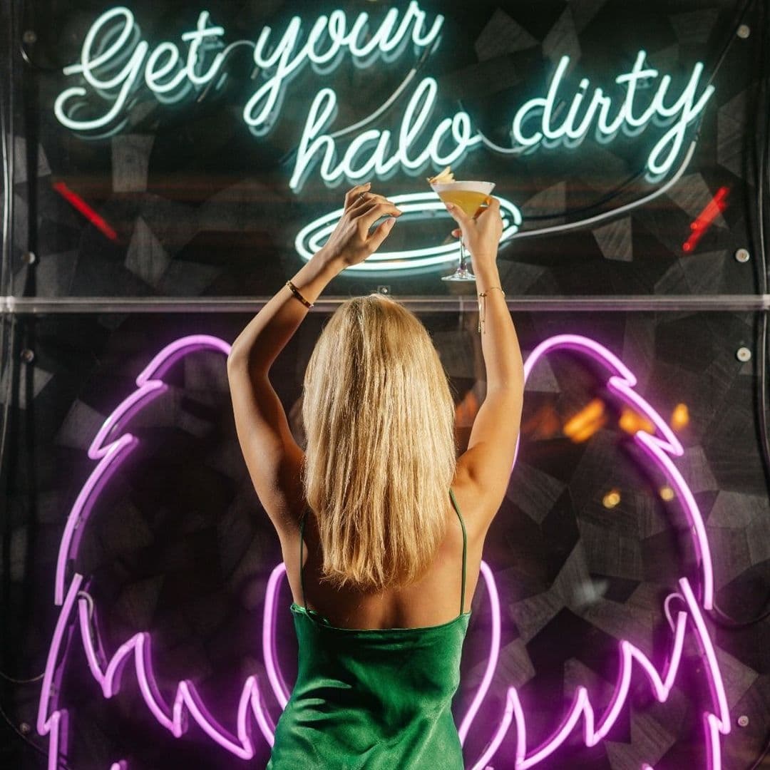 Woman in a green dress raising a martini glass under the neon sign "Get your halo dirty" at Dirty Martini.