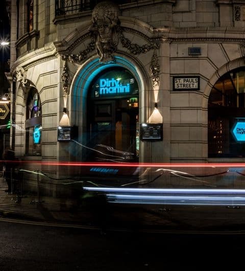 Entrance of Dirty Martini bar at night with illuminated blue signage above the door, flanked by hanging lanterns, and a light trail from a passing vehicle in the foreground.
