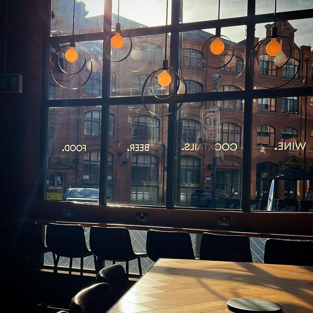 Interior view of Farrands Bar with sunlight streaming through large windows, highlighting empty wooden tables and chairs, and pendant lights with a cityscape backdrop.