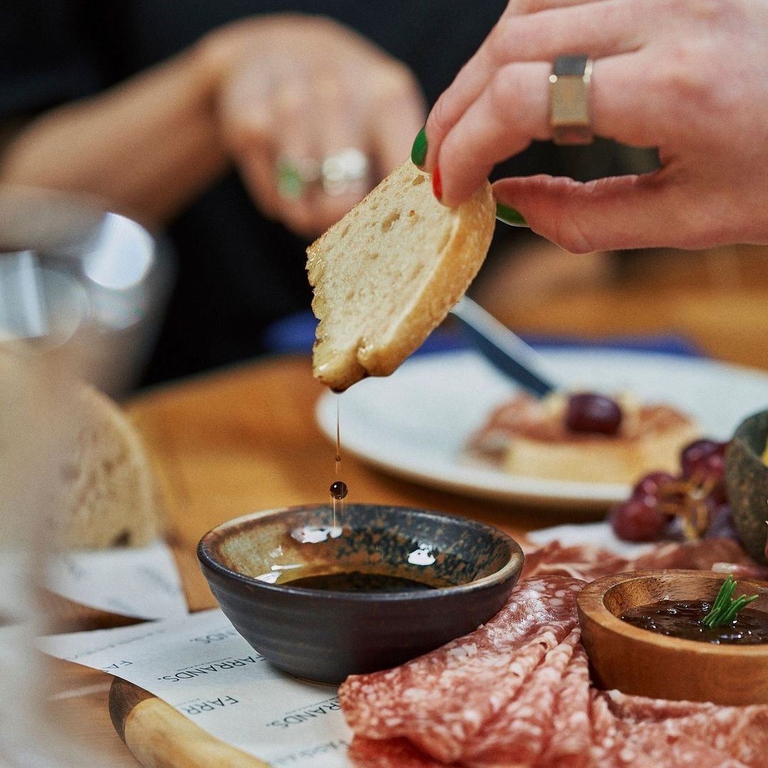 Close-up of a hand dipping bread into a bowl of olive oil at Farrands Bar, with a charcuterie platter in the background.