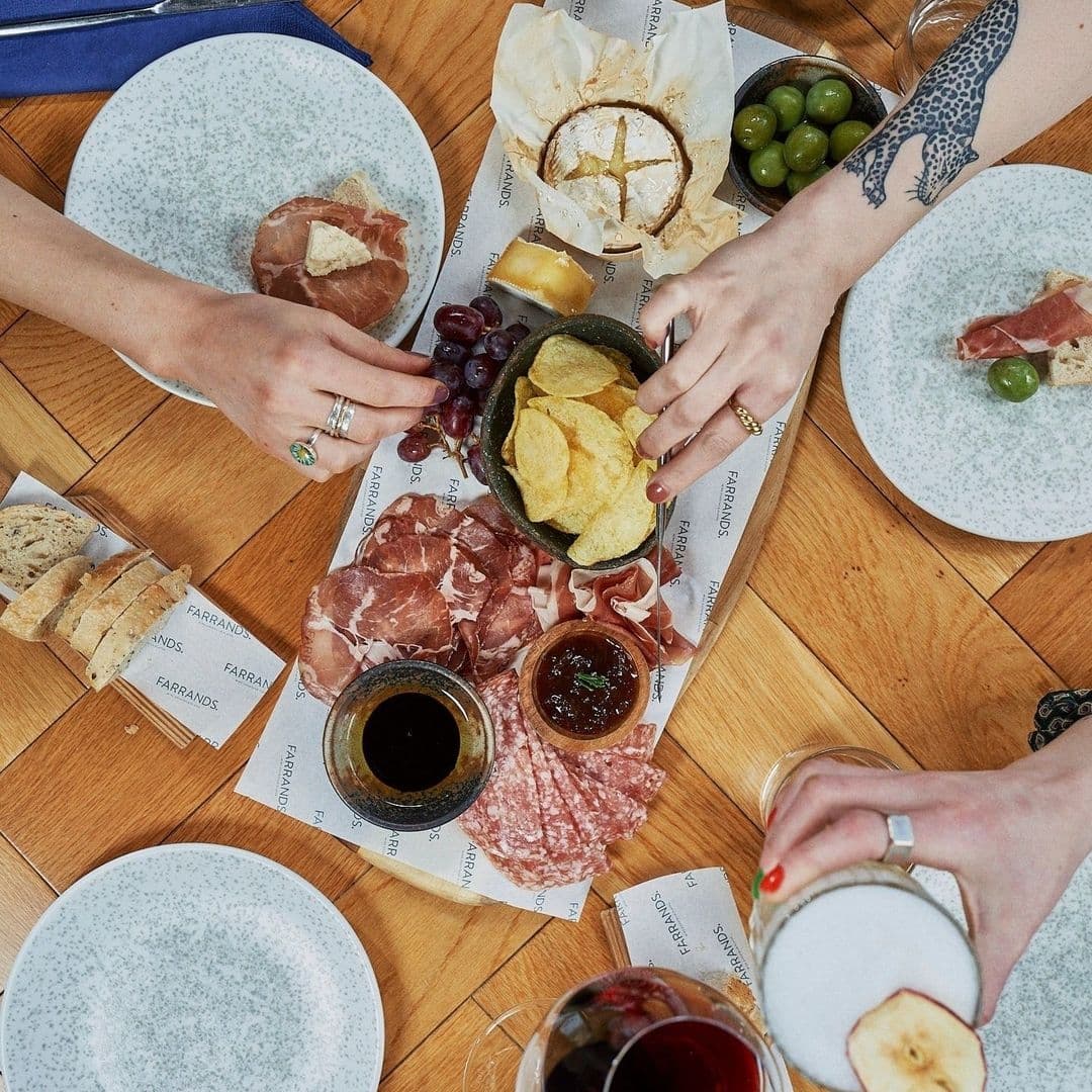 Overhead view of a gourmet charcuterie board at Farrands Bar, featuring an assortment of cured meats, cheeses, olives, and crisps, with patrons enjoying wine and beer.